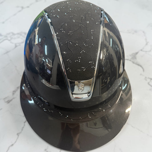 A glossy black equestrian riding helmet with a matching visor, featuring a textured black top panel with a constellation pattern and chrome accents, resting on a white marble surface.