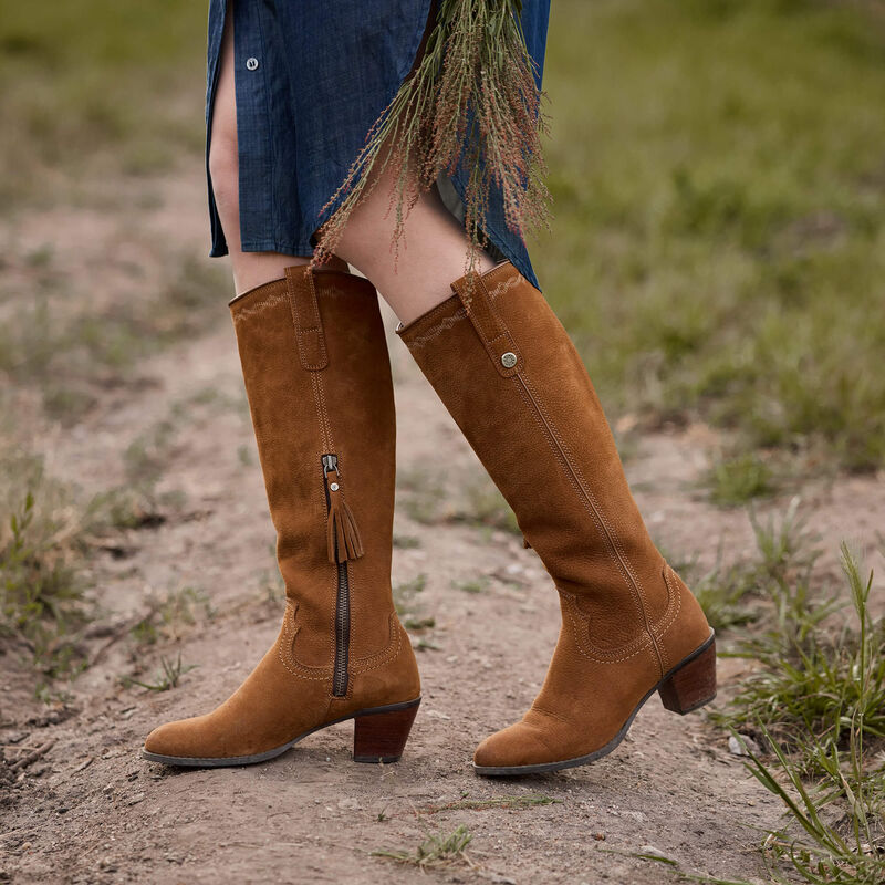 A person wearing tall tan suede knee-high boots with side zippers and tassel details, standing on a dirt path outdoors.