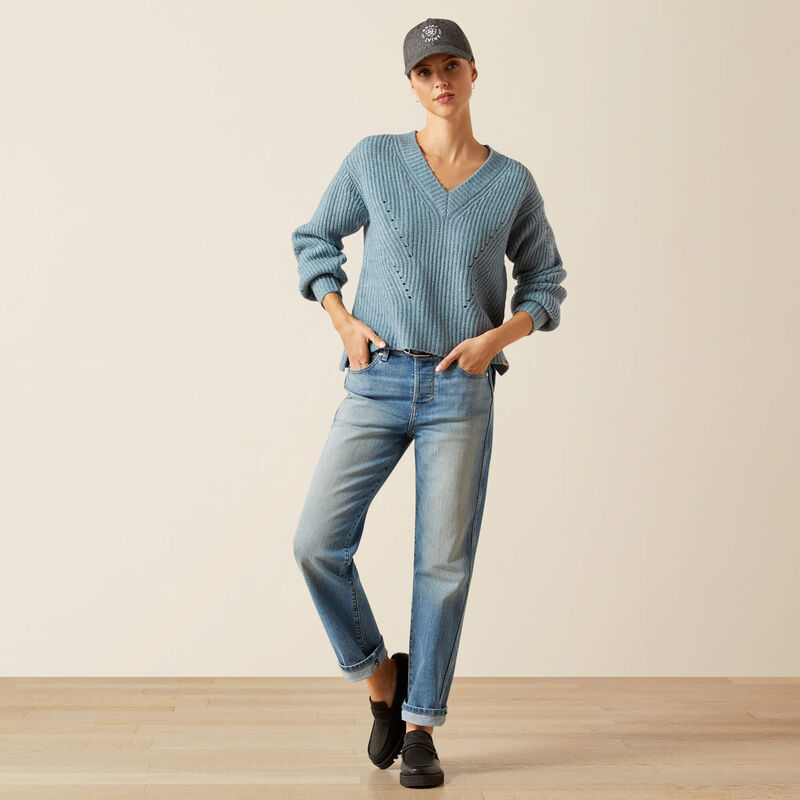 Person wearing a blue knitted sweater and jeans with a cap indoors.