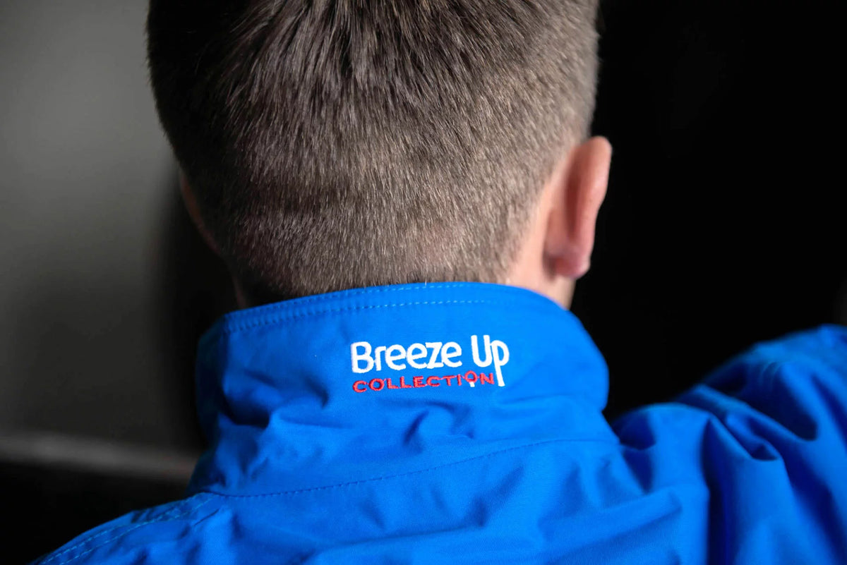 Close-up back view of a person wearing a bright blue jacket, showing the inside of the collar with the embroidered text 'Breeze Up COLLECTION' in white and red.