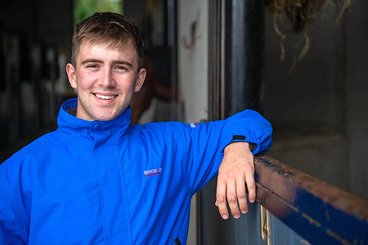 A smiling young man wearing a bright blue hooded jacket leans against a wooden railing in a stable or barn interior.