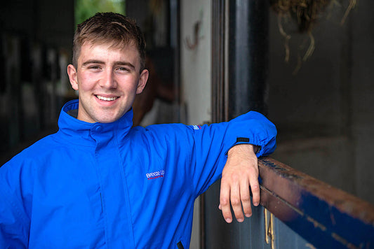 A smiling young man wearing a bright blue hooded jacket leans against a wooden railing in a stable or barn interior.