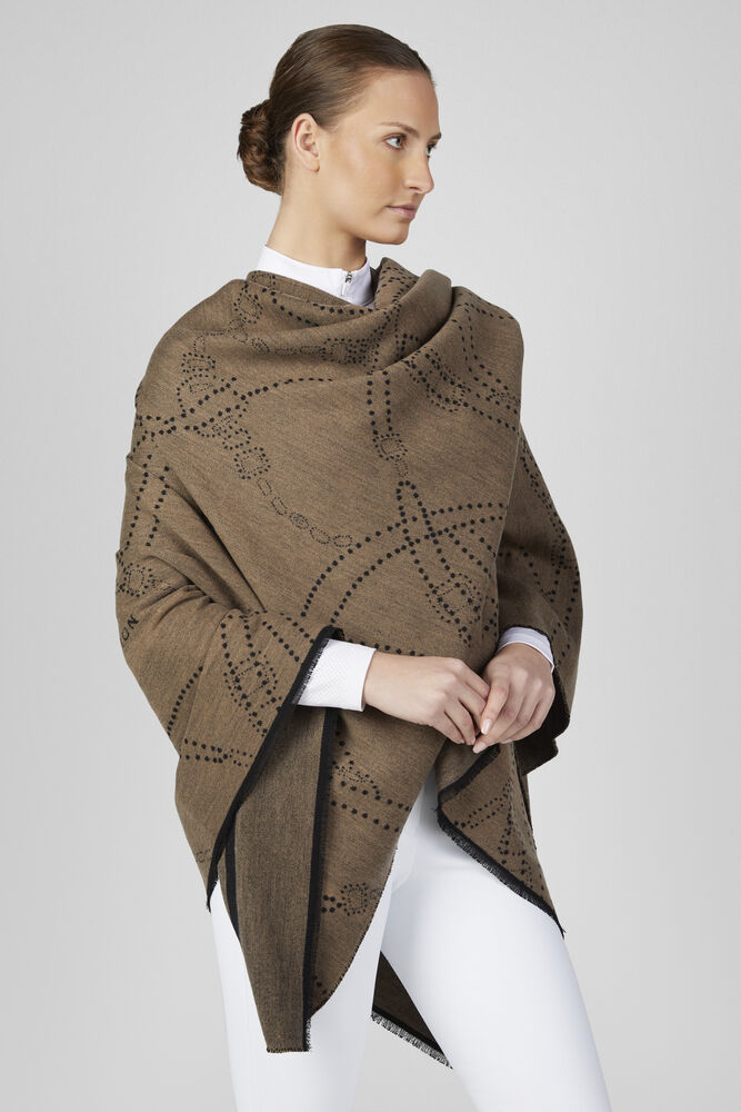 A woman wearing a brown knit poncho with black dotted geometric patterns and black trim, styled over a white collared shirt and white pants, against a light gray studio background.