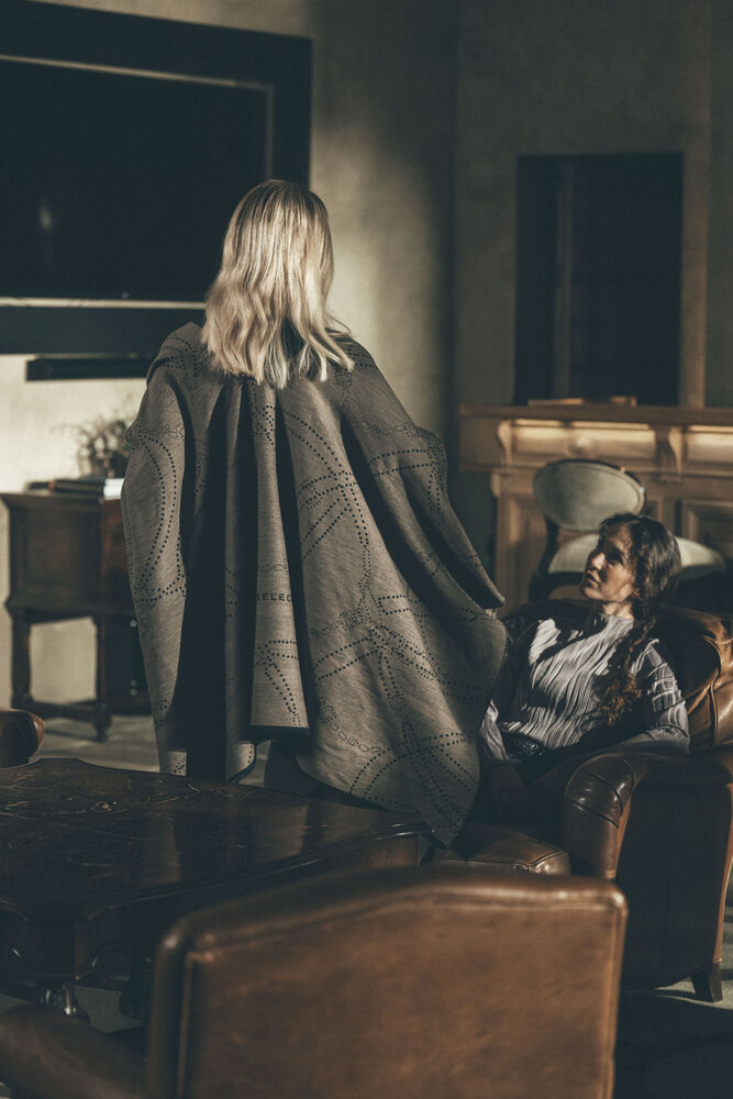 A woman with blonde hair stands with her back to the camera, draped in a large gray patterned blanket or shawl, in a dimly lit living room. Another person is seated in a leather armchair to the right.