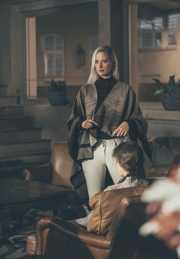 A woman stands in a dimly lit indoor room wearing a patterned brown shawl over a black top and white pants, holding a black handbag. A child is seated in a brown leather armchair in the foreground.
