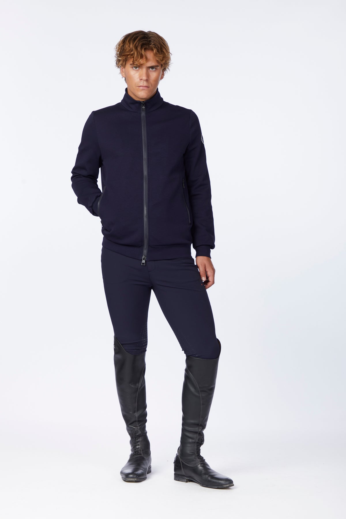 A male model wearing a dark navy blue zip-up jacket, matching slim-fit pants, and black riding boots, standing against a plain white studio background.