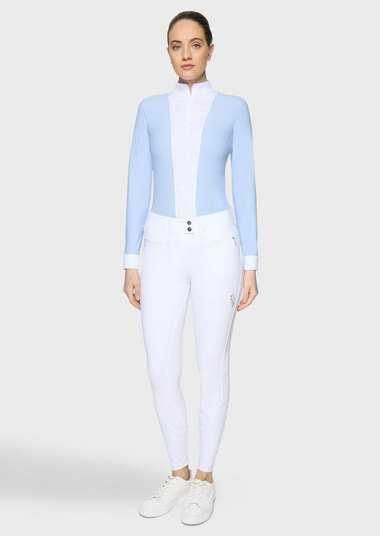 A woman modeling a light blue and white long-sleeve athletic top paired with white riding breeches and white sneakers, standing against a light gray studio background.