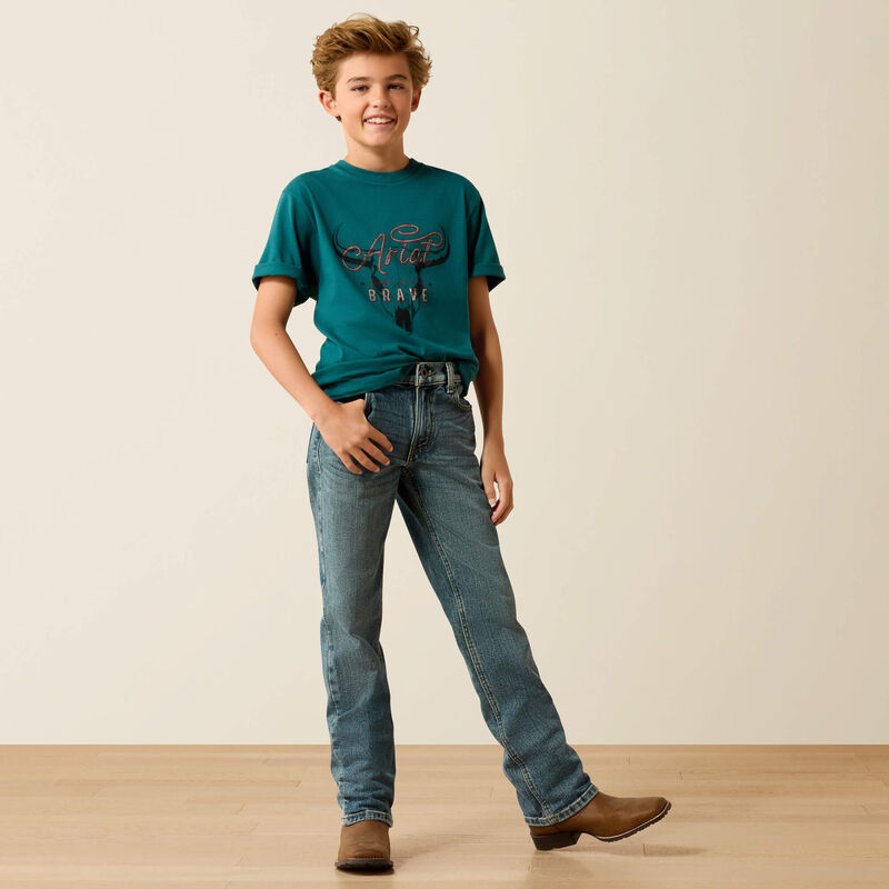 Child wearing a teal t-shirt and blue jeans standing on a wooden floor with a beige wall background
