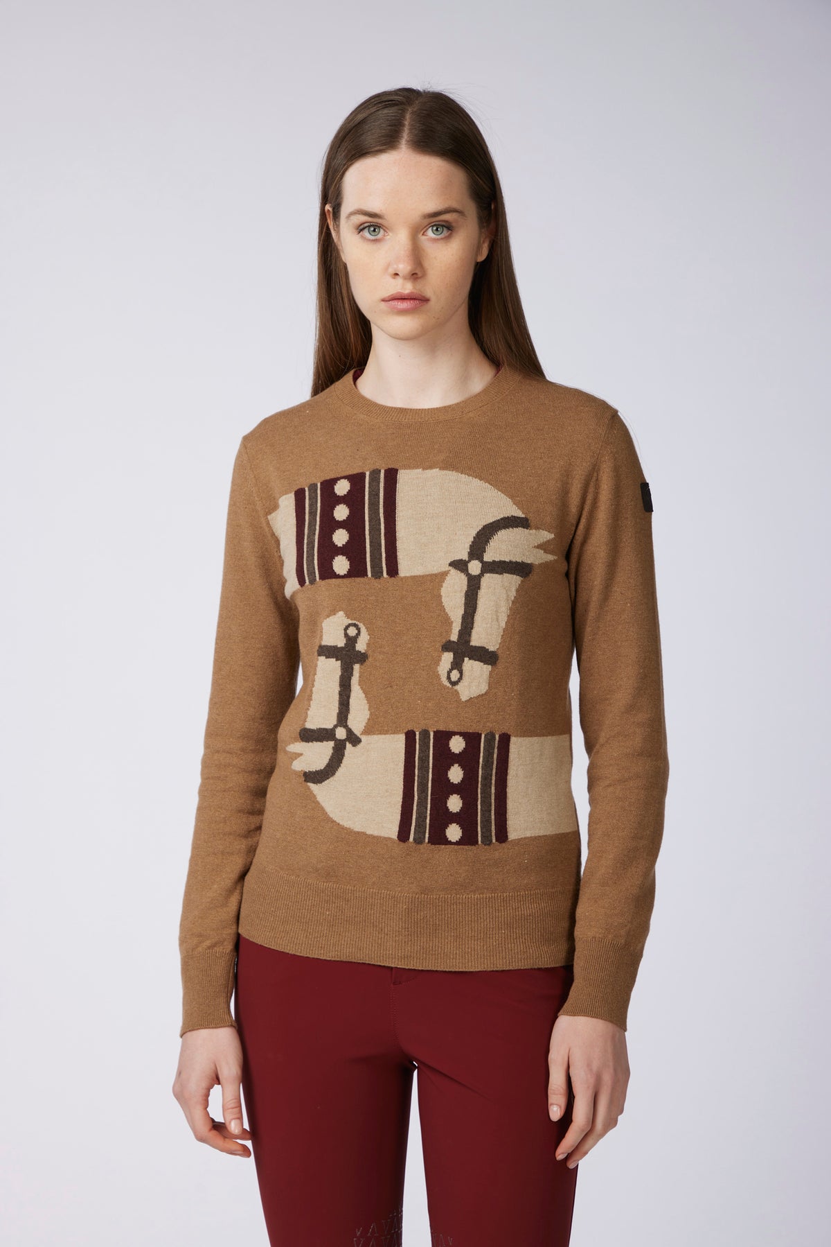 A woman modeling a brown crewneck sweater with a large graphic patch design on the front featuring beige, dark brown, and white elements, paired with burgundy pants, against a light gray studio background.