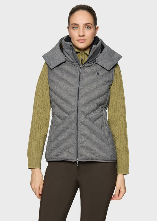 A woman modeling a gray quilted hooded puffer vest over an olive green cable-knit sweater and dark brown pants, against a light gray studio background.