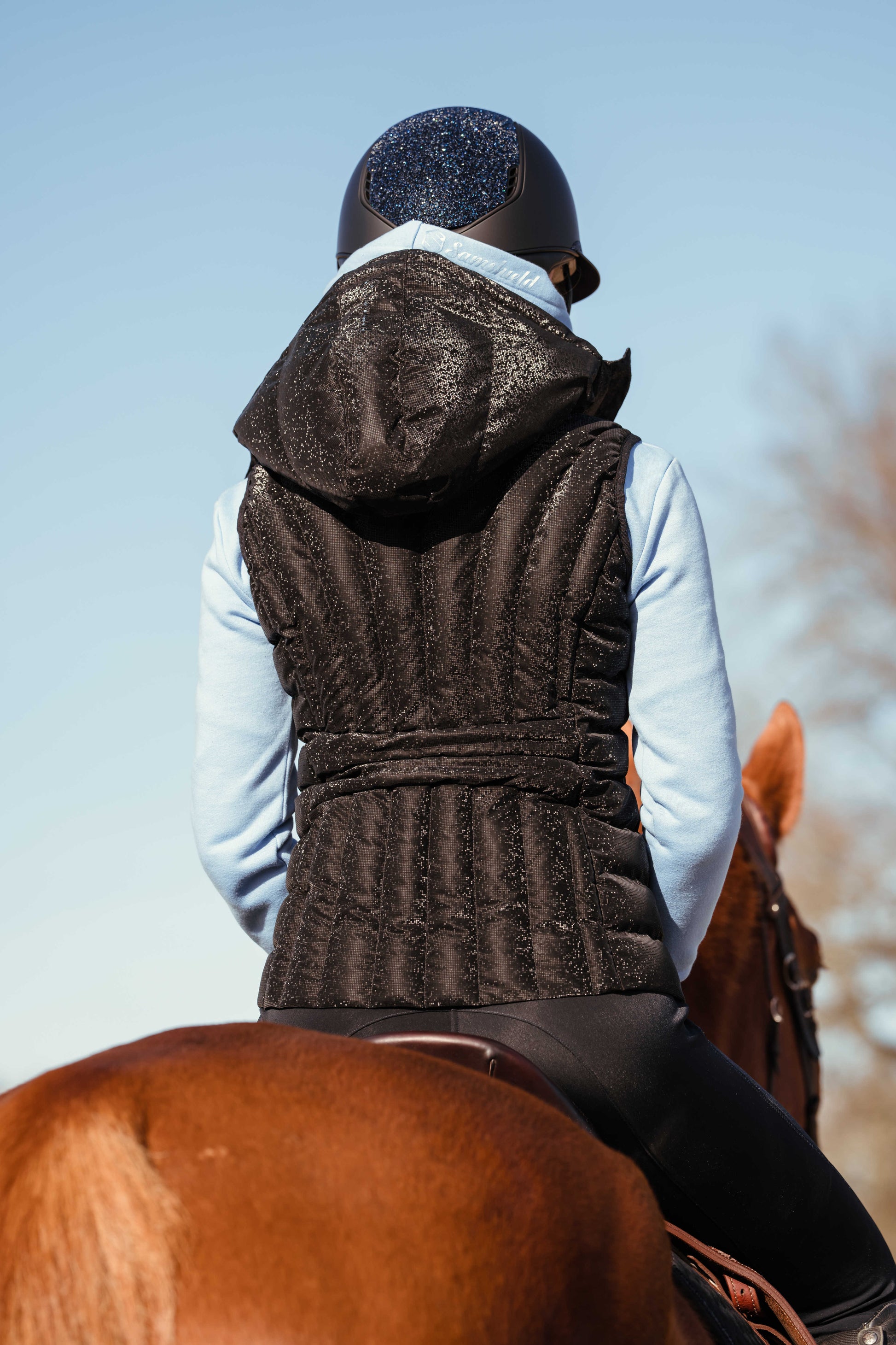The back of a woman sitting on a horse wearing a black sparkly gilet and a blue jumper with the sky as the background 