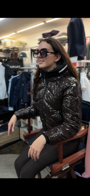 A woman wearing a shiny dark brown puffer jacket and black pants sits in a wooden chair inside a clothing store.