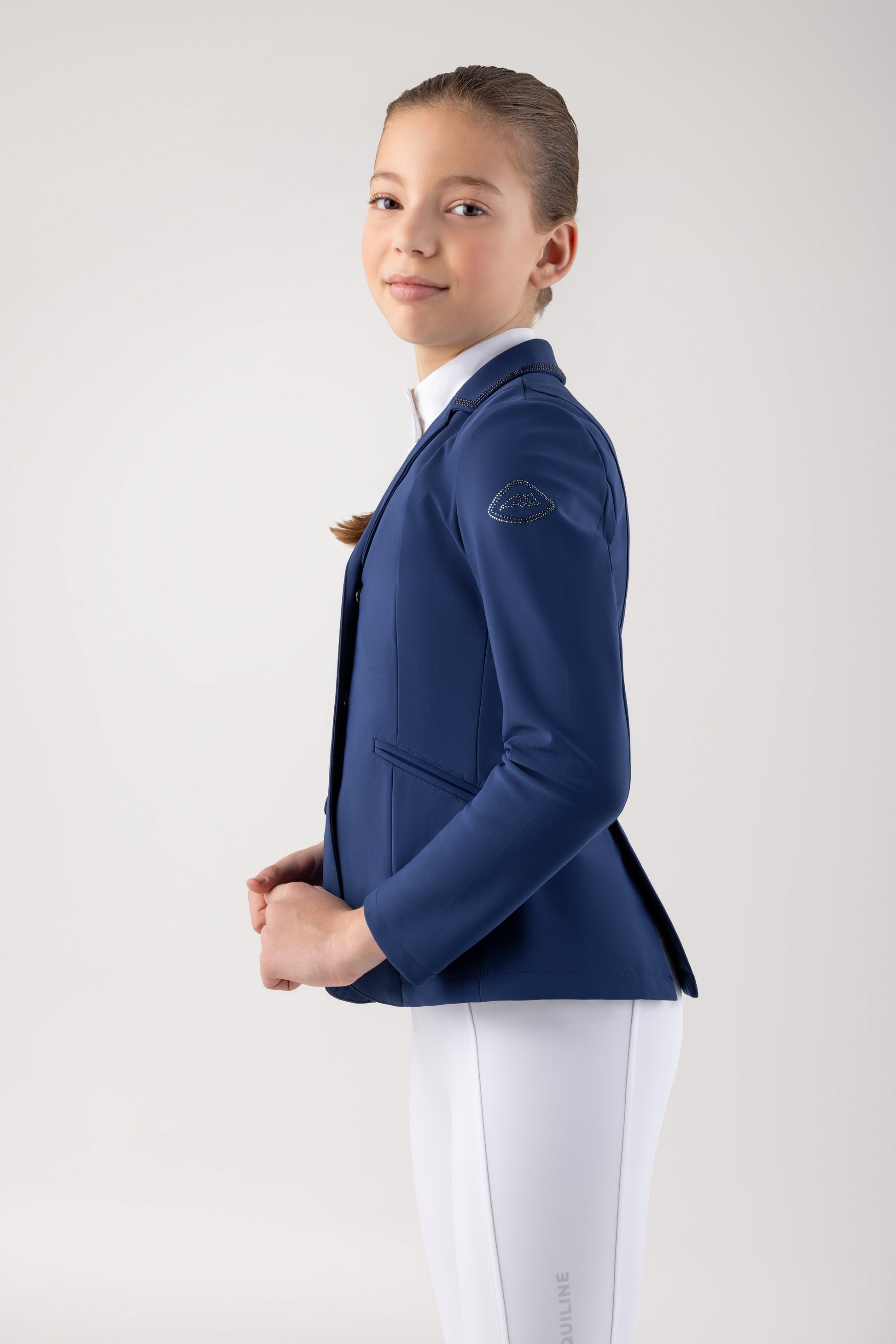 Young girl wearing a blue equestrian jacket and white pants on a plain background