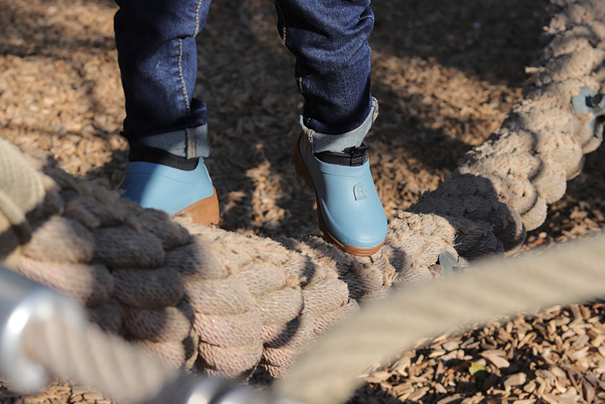 Person wearing blue rain boots stepping onto a rope bridge over a natural landscape.