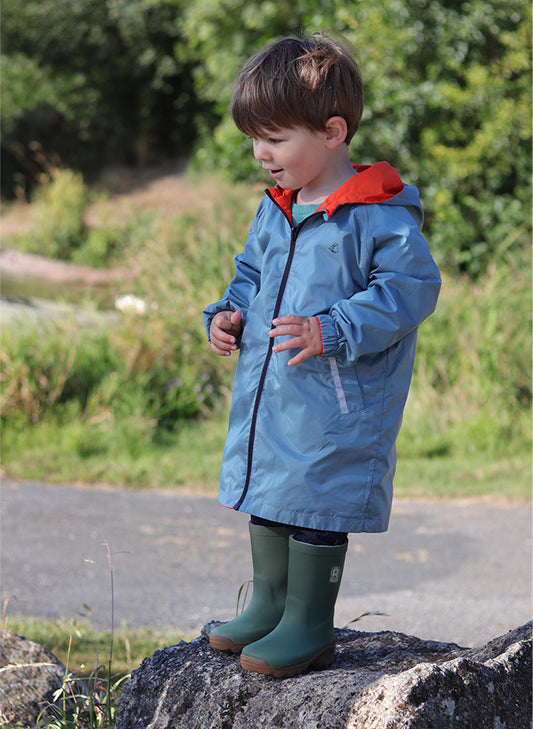 Child wearing a blue raincoat with a red hood and green boots standing on a rock outdoors.