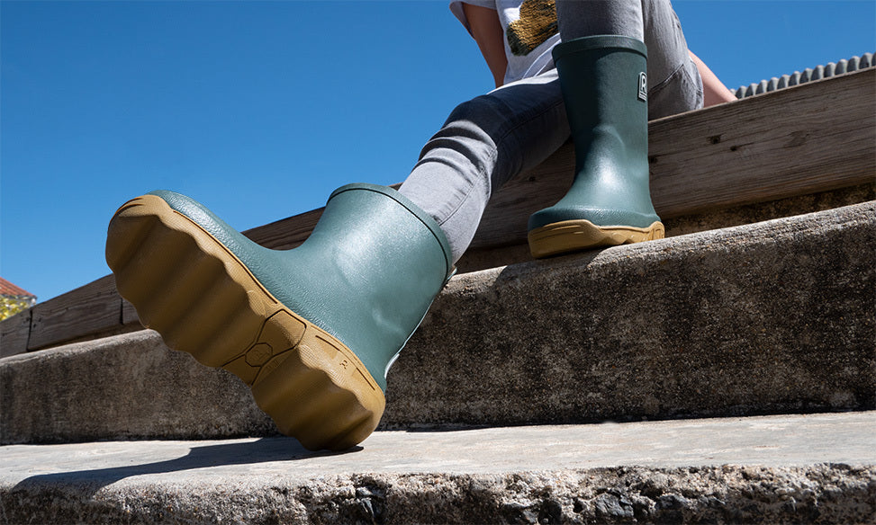 Person wearing green rubber boots with brown soles sitting on stone steps against a blue sky.