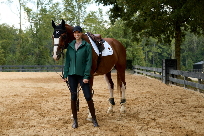 Woman in equestrian attire standing next to a brown horse in an outdoor setting with trees in the background.