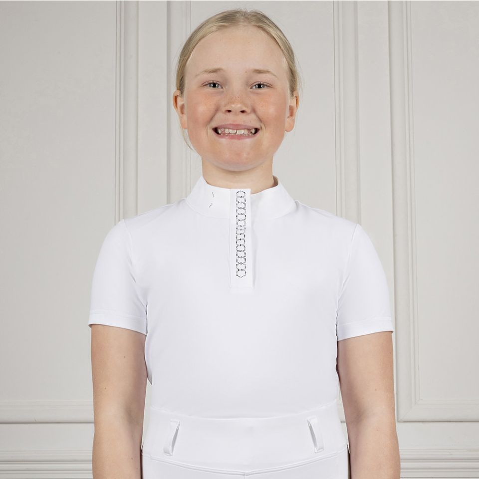 A young girl with blonde hair smiling, wearing a white short-sleeve equestrian riding shirt with a high stand-up collar and a decorative silver chain detail down the center front, set against a light gray paneled wall.