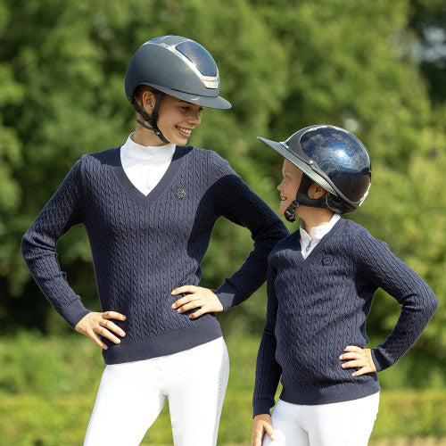 Two people wearing navy blue sweaters and white pants standing outdoors with greenery in the background.