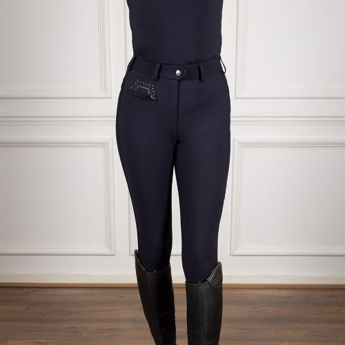 A person wearing dark navy equestrian riding breeches with a matching belt and silver snap button, paired with tall black riding boots, standing against a white paneled wall.