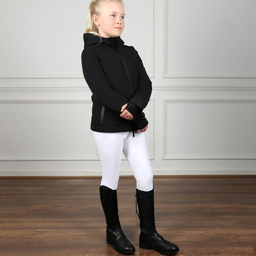 Young girl in a black equestrian jacket and white pants standing on a wooden floor.