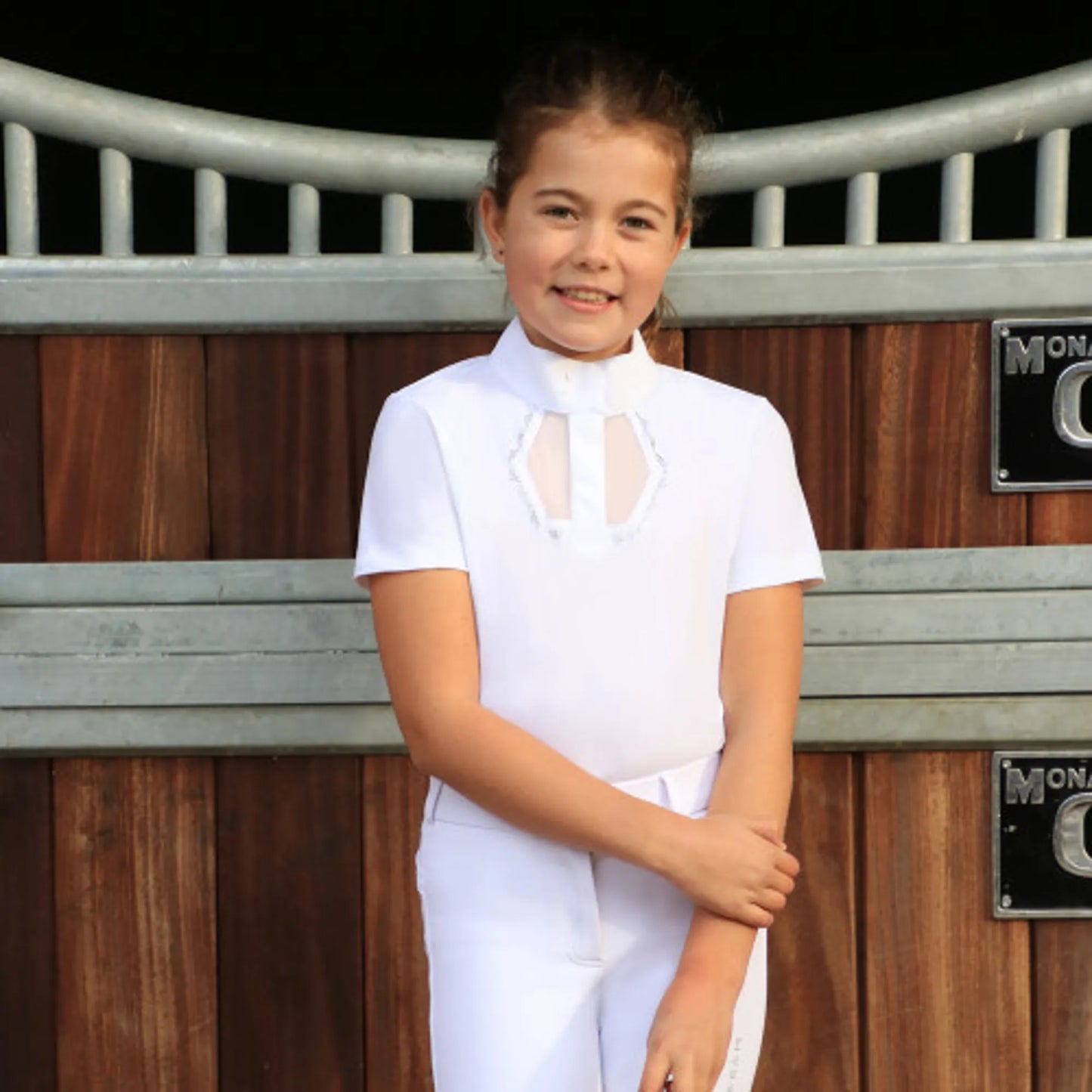 A young girl wearing a white equestrian riding shirt and matching white breeches, standing in front of a wooden stable door.