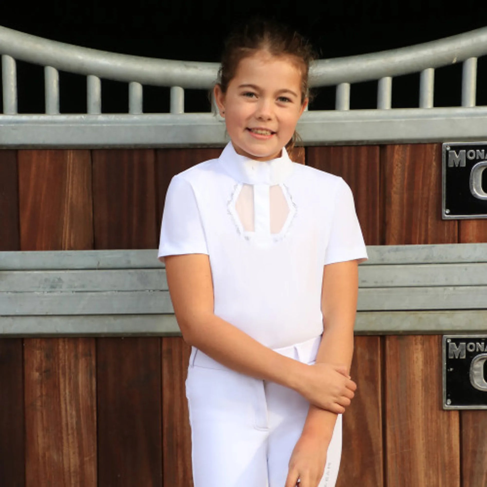 A young girl wearing a white equestrian riding shirt and matching white breeches, standing in front of a wooden stable door.