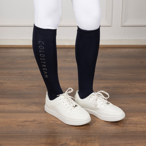 A person wearing white leggings and white sneakers with black knee-high socks featuring the brand name 'COLDSTREAM' printed vertically in white on the left sock, standing on a wooden floor.