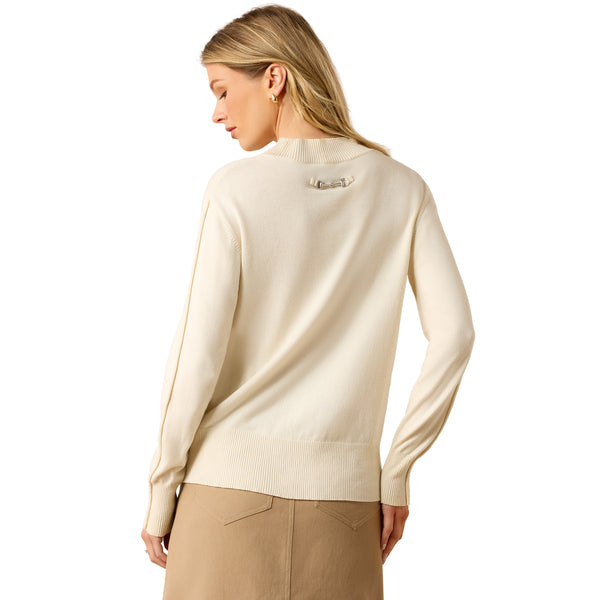 A woman shown from the back wearing a cream-colored knit sweater and beige trousers against a plain white studio background.