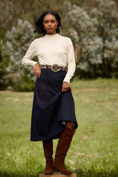 A woman standing outdoors in a grassy field wearing a cream mock-neck long-sleeve top, a navy blue high-low skirt, a patterned belt, and brown knee-high boots.