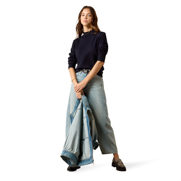 A model stands against a white studio background wearing a dark navy long-sleeve top, wide-leg light-wash blue jeans, and black lace-up shoes, while holding a folded denim jacket.