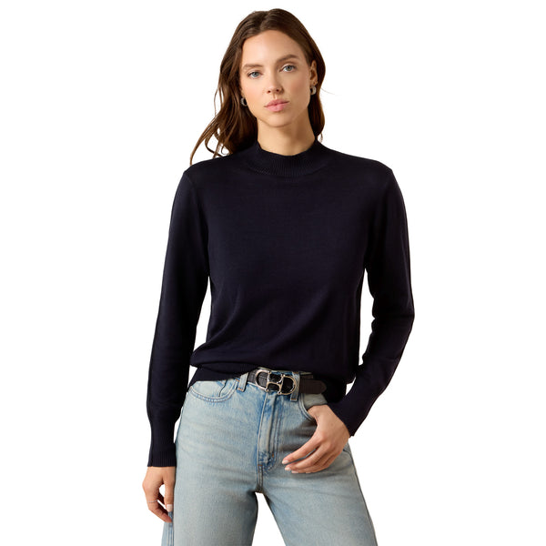A woman modeling a dark navy blue mock neck knit sweater with light-wash blue jeans and a black belt with silver hardware, shot against a solid white background.