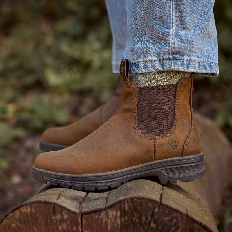 A person wearing brown suede Chelsea-style boots with elastic side panels, standing on a log outdoors. Light blue jeans are visible above the boots, and the background is a blurred natural outdoor setting.