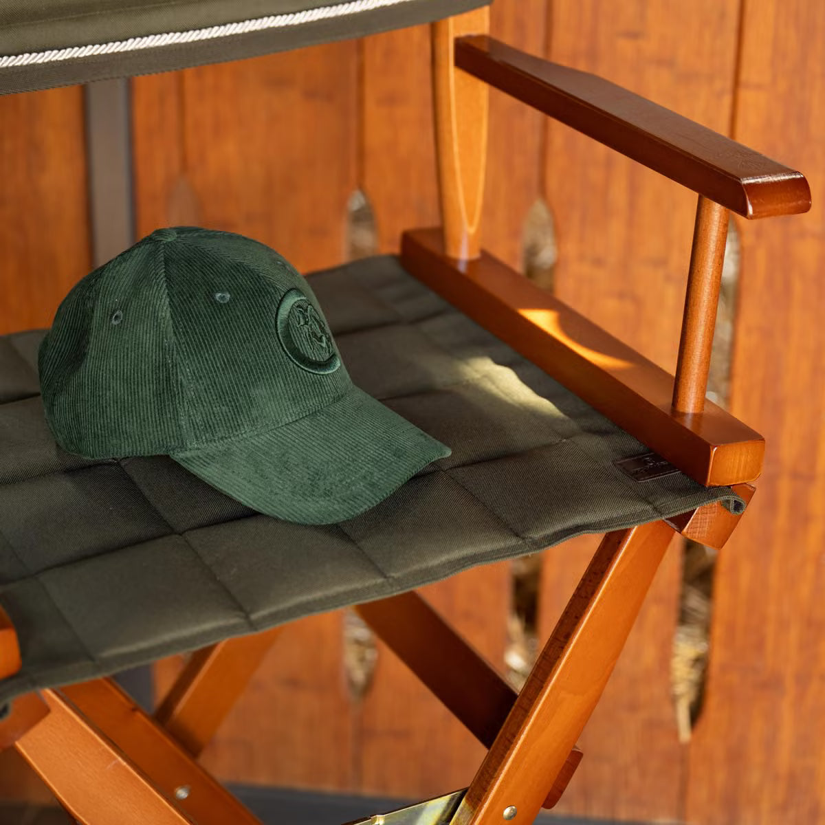 A dark green corduroy baseball cap with a circular embroidered logo sits on the seat of a wooden folding camp chair with a dark green padded cushion, set against a warm wood-paneled background.