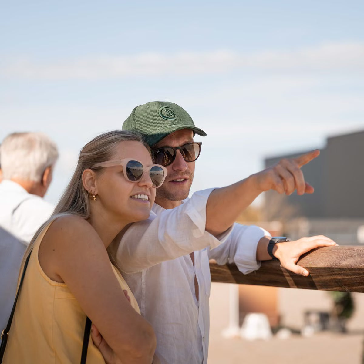A man and a woman wearing sunglasses and casual summer clothing pose together outdoors, pointing toward something in the distance under a clear blue sky.