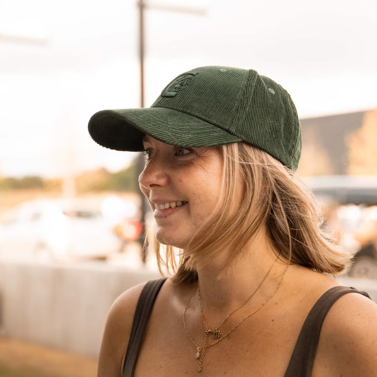 A woman wearing a dark green corduroy baseball cap with a tonal embroidered logo, photographed in an outdoor setting with a warm, blurred background.