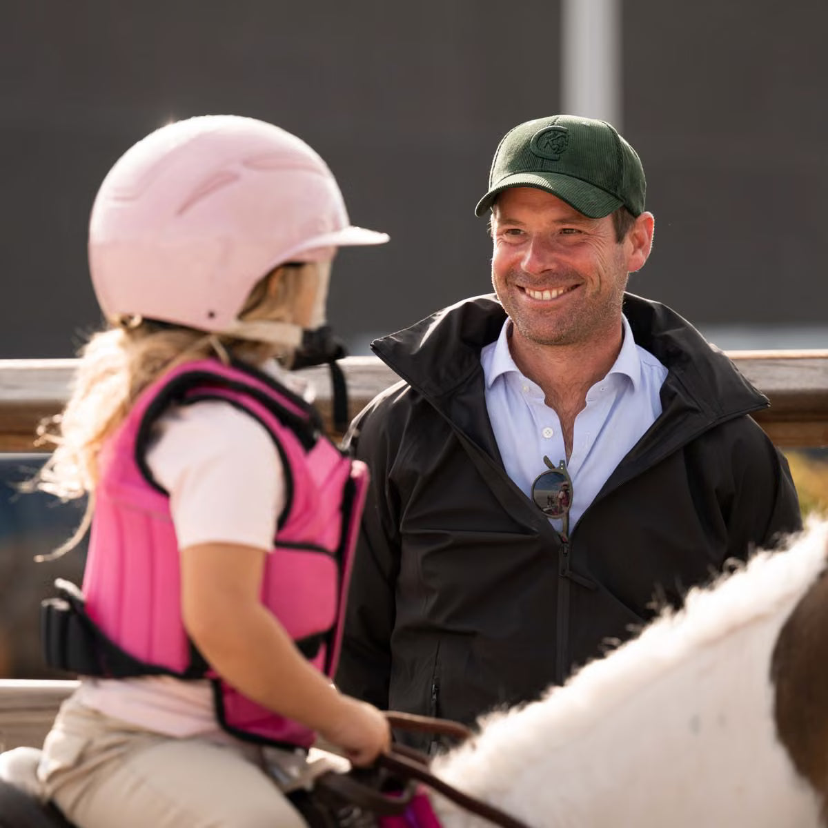 A smiling man in a green cap and black jacket sits beside a child wearing a pink riding helmet and pink protective vest, with part of a horse visible in the foreground.