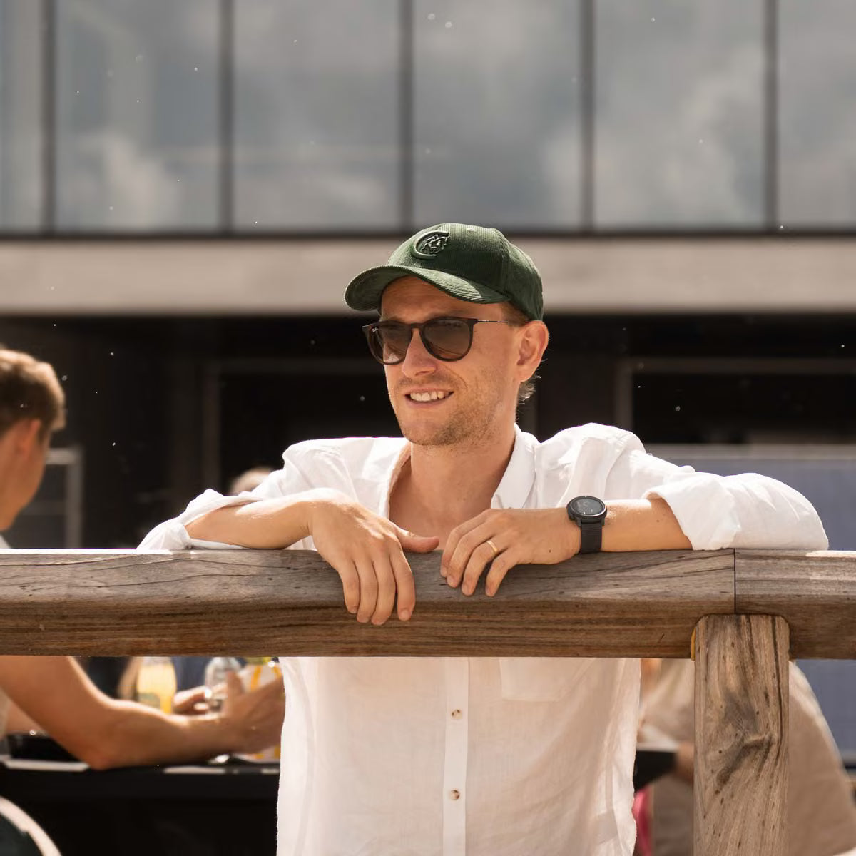 A man wearing a dark green baseball cap with a white circular logo, dark sunglasses, a white button-up shirt, and a black smartwatch leans on a wooden railing in an outdoor setting.