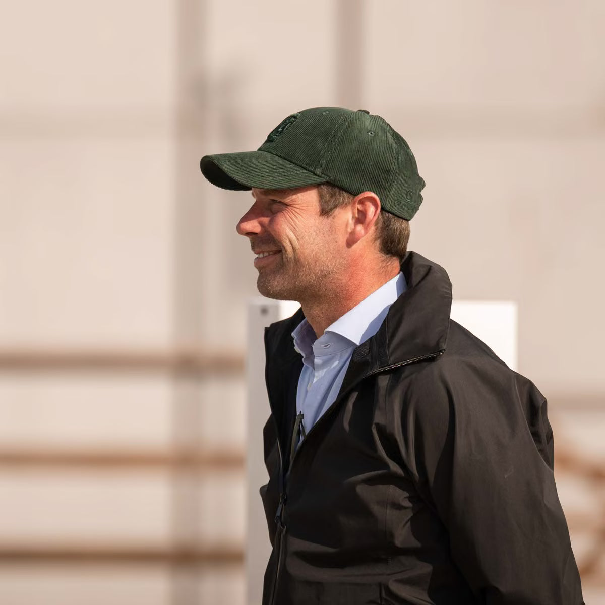 A man in profile wearing a dark green baseball cap, a black jacket, and a light blue collared shirt, photographed outdoors against a light beige wall.
