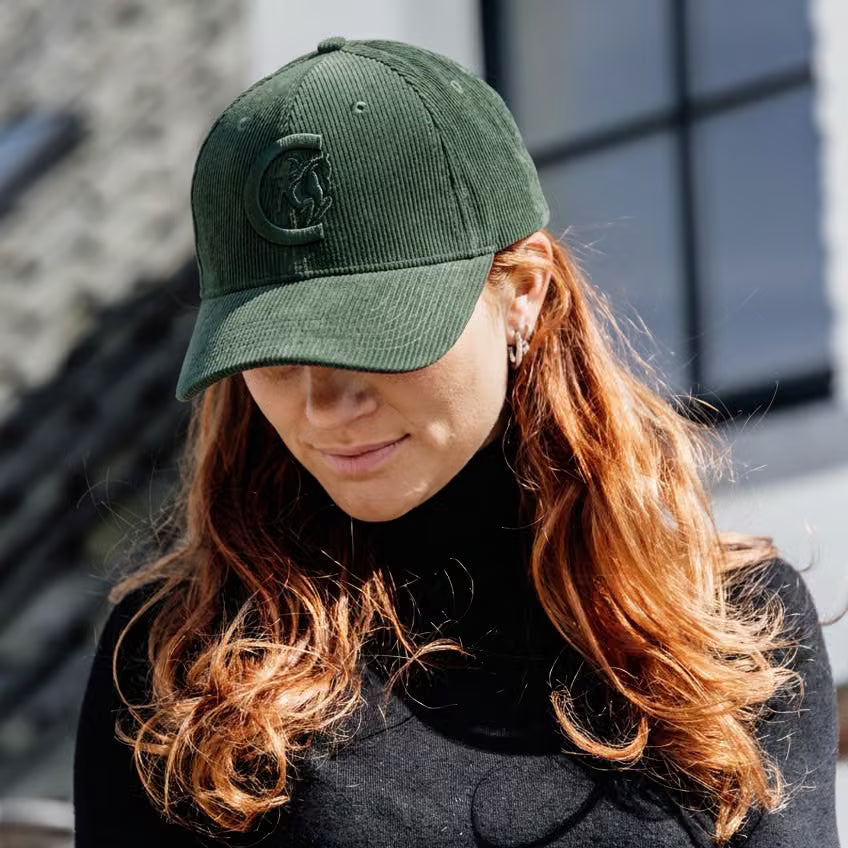 A woman with long wavy red hair wearing an olive green corduroy baseball cap with a tonal embroidered logo, photographed outdoors against a blurred building background.
