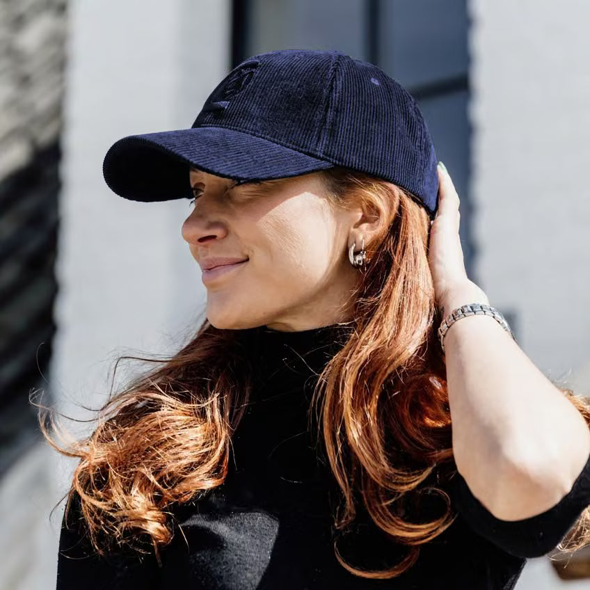 A woman with long red hair wearing a dark navy blue corduroy baseball cap and a black turtleneck, posing outdoors against a light gray building wall.