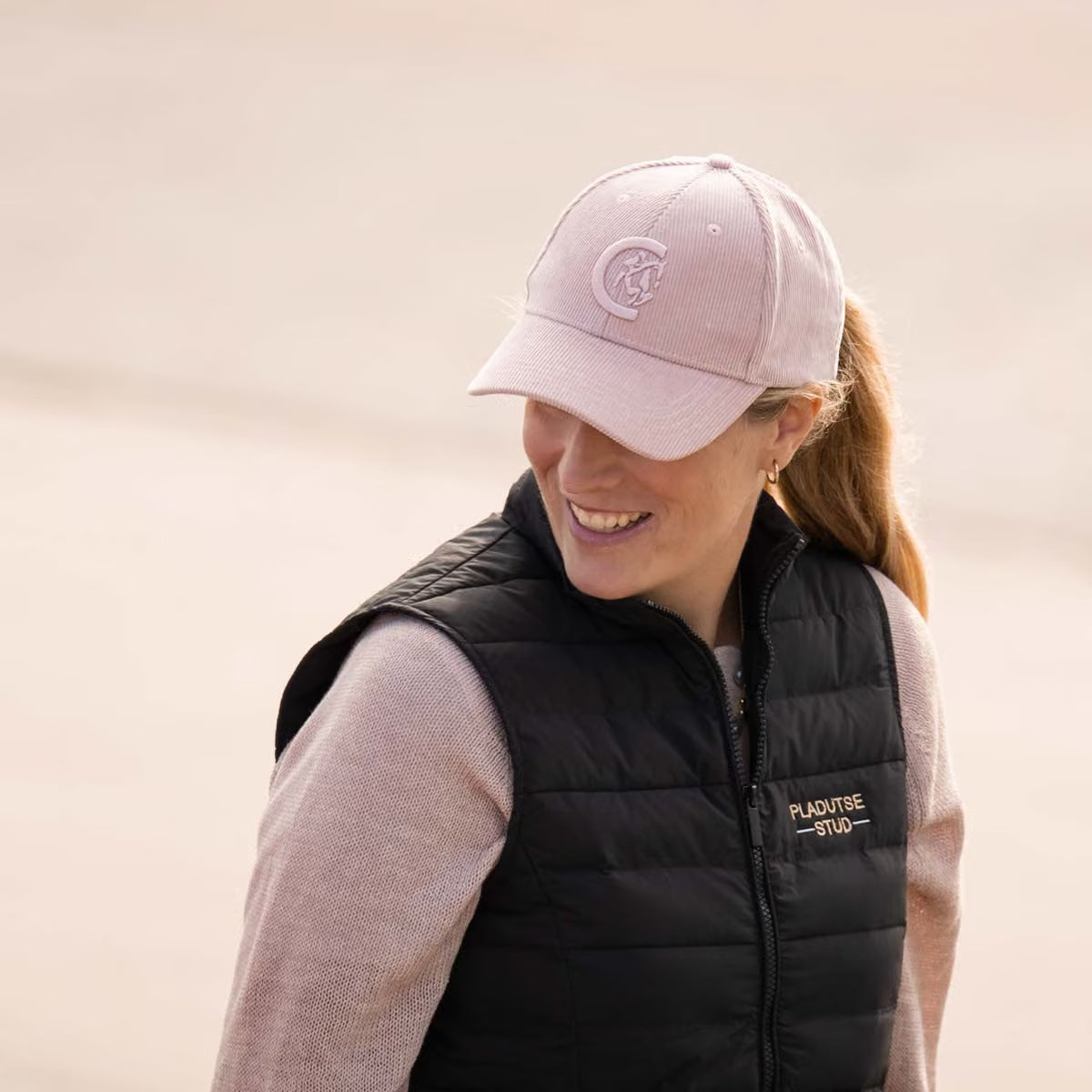 A woman wearing a light pink baseball cap with a tonal embroidered logo and a black puffer vest over a light pink long-sleeve shirt, photographed outdoors against a soft beige background.