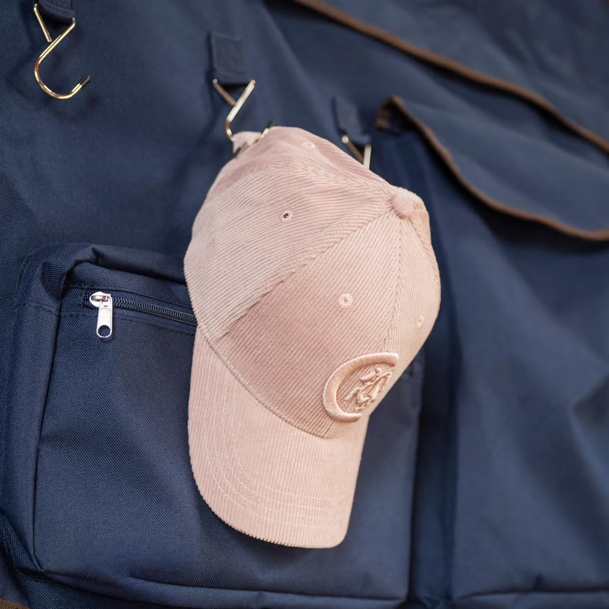 A light pink corduroy baseball cap with a tonal embroidered logo, hanging from a metal hook on a navy blue garment bag.