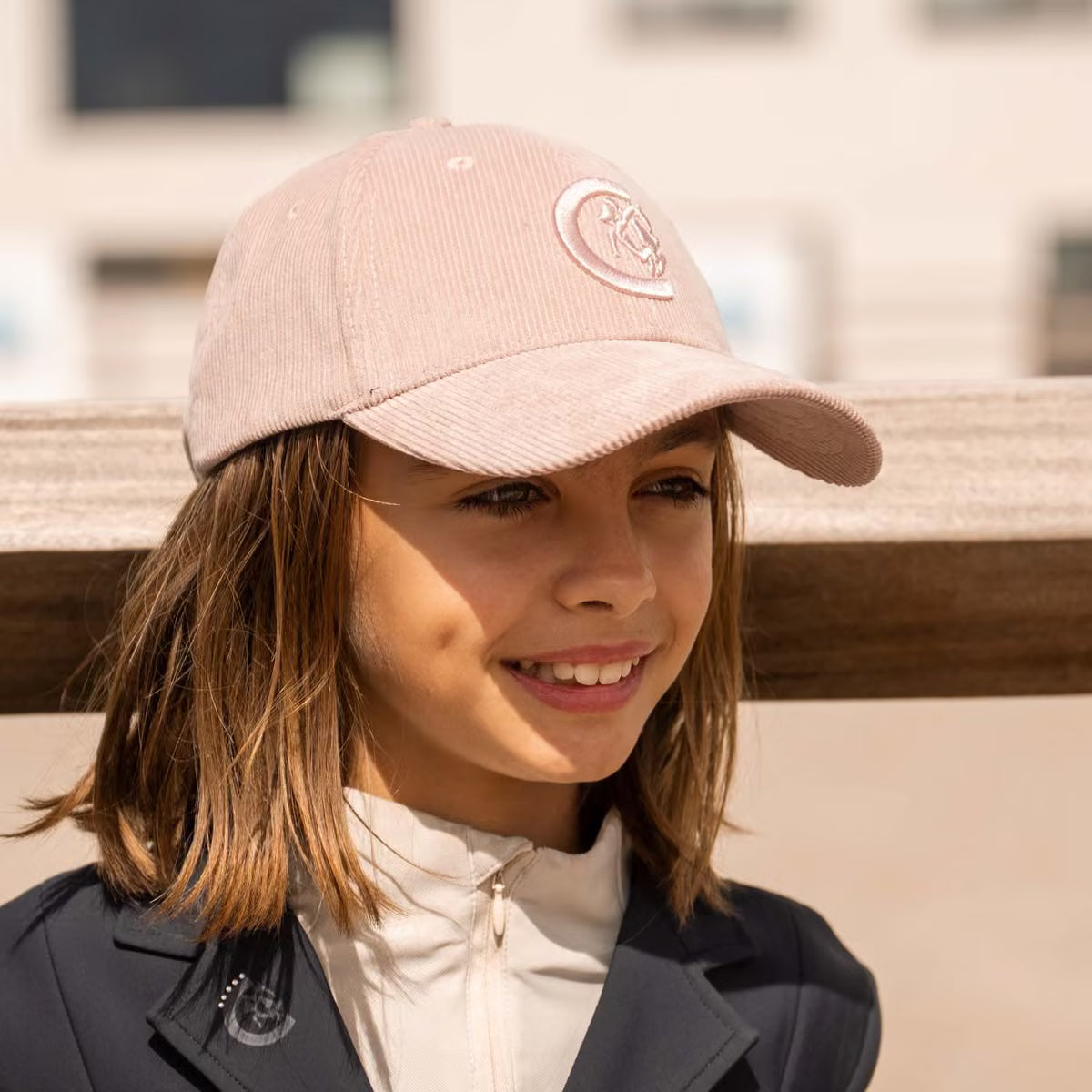 A smiling young girl wearing a dusty pink corduroy baseball cap with a white embroidered logo, paired with a white zip-up jacket and a dark navy jacket, set against a softly blurred outdoor background.