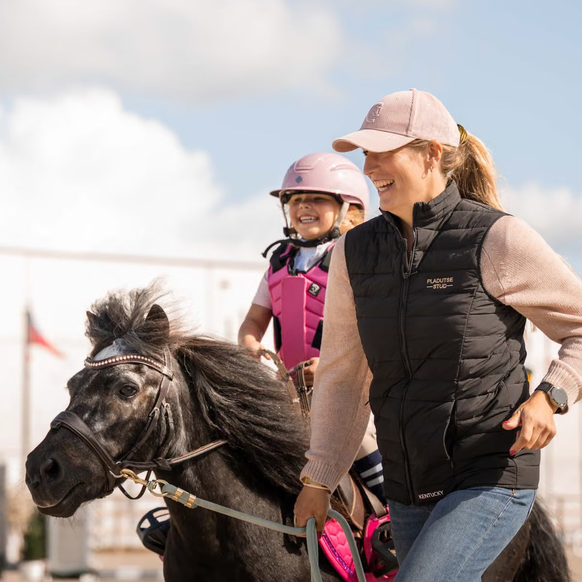 A woman and a child riding a dark horse outdoors under a cloudy sky. The woman wears a black puffer vest over a beige long-sleeve top, blue jeans, and a pink cap, while the child wears a pink riding helmet and protective vest.