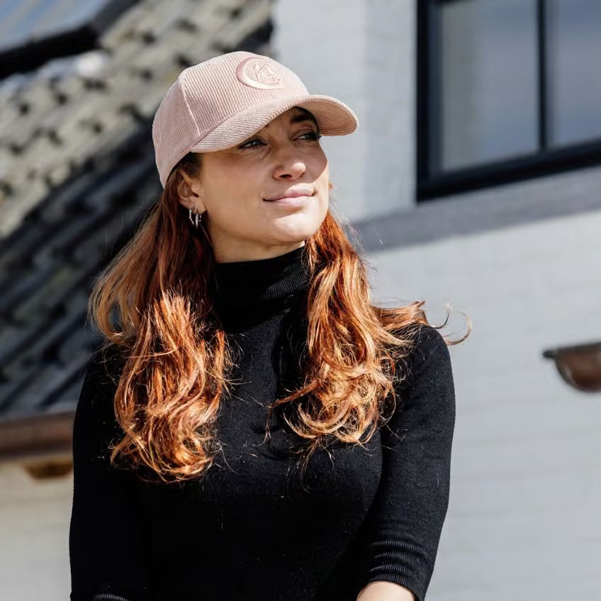A woman wearing a light pink corduroy baseball cap with a circular embroidered logo and a black turtleneck sweater, photographed outdoors against a light-colored building wall.