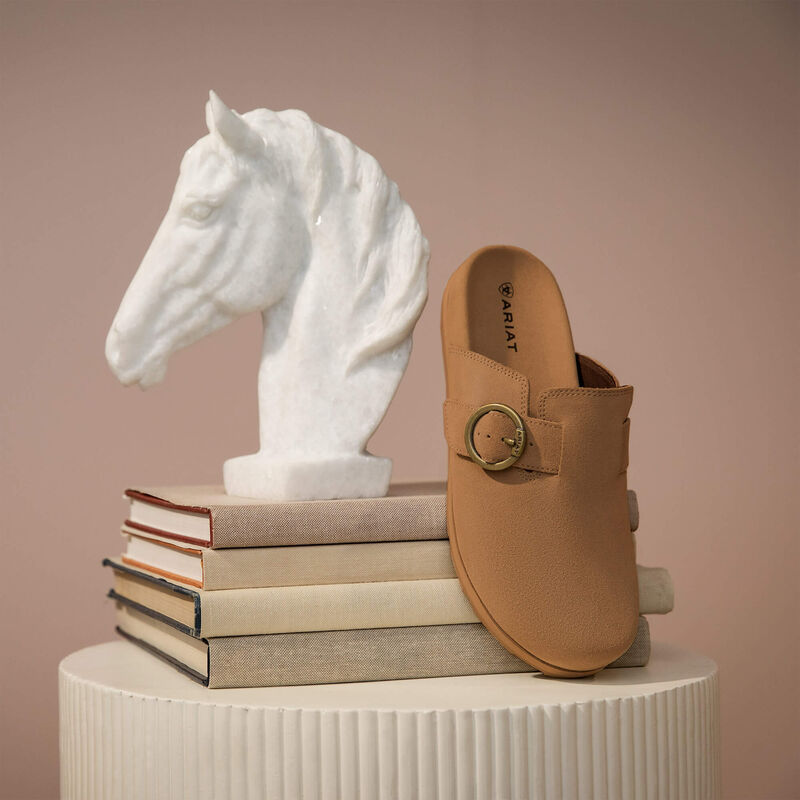 Tan shoe with gold buckle on a white pedestal next to a white horse head sculpture on books.