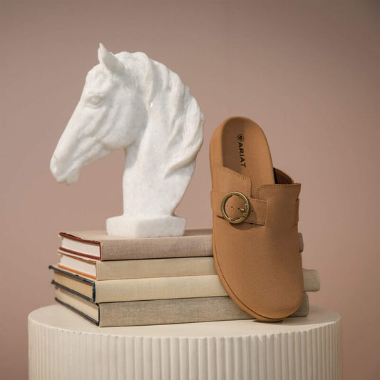 Tan shoe with gold buckle on a white pedestal next to a white horse head sculpture on books.