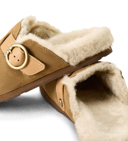 A pair of tan suede slip-on mule slippers with a soft cream shearling lining and a decorative round metal buckle, shown on a white background.