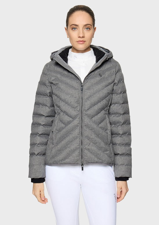 A woman modeling a gray chevron-quilted puffer jacket with a black hood lining, worn over a white top and white pants, against a light gray studio background.